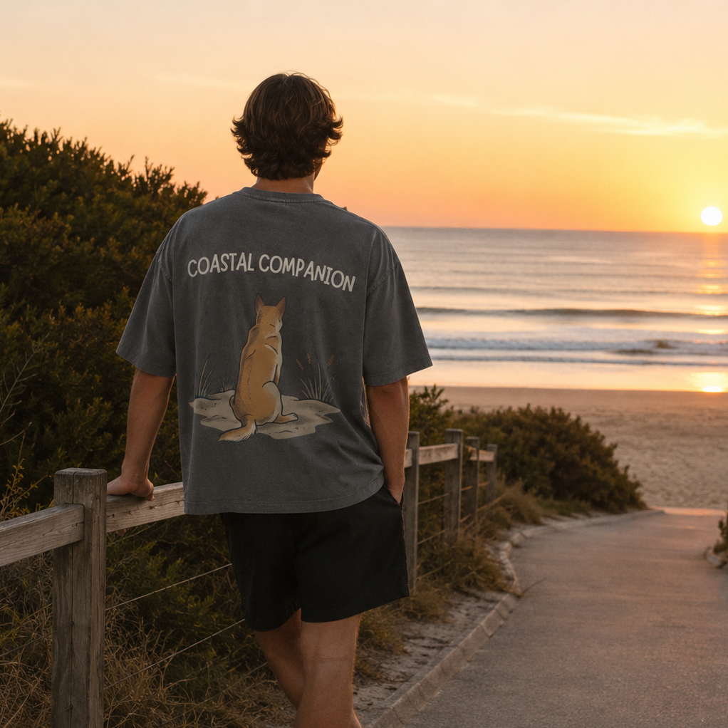 A man stands on a beach path at sunset wearing a dark gray t-shirt with 'COASTAL COMPANION' and a cat graphic printed on the back, paired with black shorts.